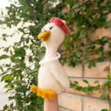 A handmade wool felt duck finger puppet, from the 'Honeymoon' collection, is worn on a female hand against green foliage and a brick wall. This off-white felt duck has a textured finish, bright yellow beak and feet. Adorned with a red felt beret and a string of white pearls, this charming handcrafted felt kids toy reflects its delightful honeymoon attire.