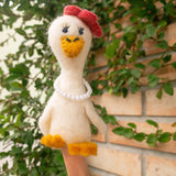 A charming white wool felt duck finger puppet is worn on a person's finger, positioned against a backdrop of green leaves and a rustic brick wall. This handcrafted felt duck, part of a honeymoon-themed duo, features a bright yellow beak and feet, small blue eyes, a red bow on its head, and a delicate pearl necklace.