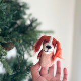 A handcrafted Cavalier King Charles Spaniel felt finger puppet is playfully held by a hand against a festive Christmas tree. This charming felt puppet for finger play features floppy orange ears, a white forehead blaze, a black nose, and a stitched smile on its light brown felt body. Its whimsical design, handcrafted from felt, sparks imaginative play.