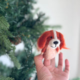 A handcrafted Cavalier King Charles Spaniel felt finger puppet is held by a female hand, set against a blurred, festive Christmas tree. This charming orange and white wool felt finger puppet, designed with a smiling face, is part of a unique set of two, showcasing its delightful craftsmanship.