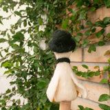 A charming white wool felt duck finger puppet from the 'Duck Couple' set sits on a person's finger against a lush green ivy wall. This handcrafted felt toy, featuring a playful black hat and scarf, is ready for interactive storytelling and imaginative play, highlighting its delightful character.