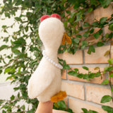 An off-white handcrafted felt duck finger puppet is playfully held by a female hand against a brick wall partially covered with green vines. This unique duck puppet, expertly crafted in Nepal, features vibrant red tufts, an orange beak and feet, and a charming white beaded necklace. Showcasing quality craftsmanship, this felt toy is perfect for imaginative storytelling.