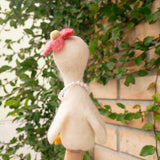 A handcrafted felt duck finger puppet, part of the 'Duck Couple on Honeymoon' set, is held by a female hand against a brick wall partially covered with green vines. This cream-colored wool felt duck puppet features a red flower atop its head and a strand of white beads around its neck. Its fuzzy texture adds to its whimsical charm.