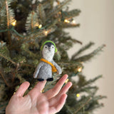 A handmade felt penguin finger puppet, part of a set of two, is held by a female hand in front of a twinkling Christmas tree. This unique needle felted penguin puppet features a gray body, black face and feet, and bright green felt headphones, bringing playful charm to any collection.