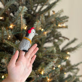 A hand holds up a handcrafted felt penguin finger puppet from a set of two, placed against a softly lit Christmas tree backdrop. This unique grey and white felt puppet for finger play is adorned with bright red headphones and a yellow scarf.