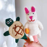 A female hand holds a set of two handcrafted felt turtle and rabbit finger puppets against a plain white backdrop. The green felt turtle finger puppet features a tan shell with a brown swirl design and small, round eyes. Beside it, the white felt rabbit finger puppet has pink inner ears, a pink nose, and a red mouth.