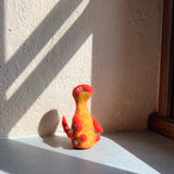 A vibrant wool felt Brontosaurus finger puppet from the 'Dino Brothers' set rests on a gray surface, bathed in natural light against a textured wall. This handcrafted needle-felted toy has a lifelike 3D design, featuring a long neck and a tail. Its body transitions from red at the head to yellow on the main body, complemented by red arms and white spots. This unique felt toy for kids is ideal for imaginative play.