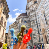 A set of three handcrafted wool felt Brontosaurus finger puppets are playfully showcased on fingers, set against a backdrop of classical urban architecture under a bright sky. These unique Brontosaurus puppets come in blue, green, and red, each adorned with white spots. Their handcrafted nature emphasizes a whimsical blend of playful toys and iconic urban scenery.
