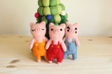 A set of four handmade felt finger puppets, depicting the Three Little Pigs and the Big Wolf, are playfully arranged on a light wooden surface against an off-white background. Each unique pig felt finger puppet features distinct facial details, with one having closed eyes, another black spots, and the third open eyes. The pigs wear colorful orange, red, or blue overalls, adding charm to these handcrafted felt kids toys.