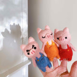 Three handmade felt pig finger puppets from a Three Little Pigs story set are held on a female hand, presented against a white wall with a faint shadow. These unique felt pig finger puppets, crafted from soft pink wool felt, feature pointy ears and distinct pink snouts. One pig wears blue overalls with a grumpy expression, another orange with closed eyes, and the third red, encouraging imaginative play.