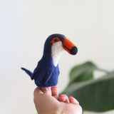 A hand holds a small, colorful felt bird finger puppet against a plain background. The puppet has a blue body with a red beak and feet, featuring simple, stylized design without textures or patterns. This handmade wool felt bird finger puppet is part of a set of 5 delightful designs from Ganapati Crafts Co., expertly crafted in Nepal for creative play and storytelling.