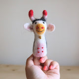 A handcrafted 100% wool felt cow finger puppet is held gently by a female hand with bright pink nails, set against a simple light beige wall and light-colored wooden surface. This soft, textured felt farm animal finger puppet features a white face, smiling orange nose, black and white hair, red-tipped horns, a gold bell, and pink spots, showcasing its unique design.