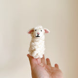 A charming white wool felt sheep finger puppet, part of a farm animal set, is held upright by a female hand against a plain backdrop. This handcrafted sheep finger puppet features a textured, soft body with pink inner ears, a pink nose, and black eyes, showcasing its detailed craftsmanship and gentle playfulness.