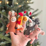 Five charming, handmade wool felt farm animal finger puppets are presented together by a female hand against a softly blurred Christmas tree background. The collection features a brown horse, a yellow and orange hen, a white and black cow with a small golden bell, a grey sheep, and a pink pig. Each handcrafted felt finger puppet displays stitched details for their smiling faces and unique features.