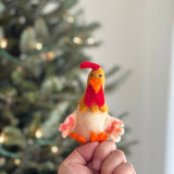 This festive, handmade 100% wool felt hen finger puppet from the Farm Animals set is held playfully in a hand, with a blurred, warmly lit Christmas tree in the background. The detailed hen puppet features a golden yellow head with a red crest and wattle, a pointy orange beak, and beady eyes. Its soft, off-white body has red and orange-tipped wings, complementing its orange feet.