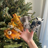 A set of five handcrafted felt safari animal finger puppets is presented by a female hand against a festive Christmas tree background. The collection features a felt lion finger puppet with a golden mane, a spotted felt cheetah finger puppet, a long-necked felt giraffe finger puppet, a gray felt elephant finger puppet, and a striped felt zebra finger puppet. These unique, fair-trade felt kids toys are ideal for imaginative play.
