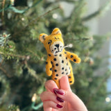 A handmade felt cheetah finger puppet is held by a female hand with dark red nails, against a blurred Christmas tree background with soft, twinkling lights. This charming yellow and black felt cheetah puppet features distinctive spots, a small smiling face, and whiskers. Crafted for playful interaction, this bendable and cuddly felt toy supports fair-trade artisans.