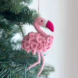 A pink felt flamingo ornament, hanging from a green Christmas tree, is set against a plain white wall, evoking a festive and cheerful mood. This handmade felt flamingo ornament features a darker pink peak and a white stripe, crafted with a textured body and suspended by a jute string.