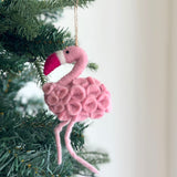 A handcrafted felt flamingo ornament hangs from a green Christmas tree branch by a jute string, with a blurred white wall in the background. The pink felt ornament is detailed with a body of small, rounded felt pieces, thin pink legs, and a dark pink and white beak, bringing whimsical charm to the holiday decor.