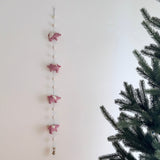 A pink felt flying pig garland is artfully displayed against a clean white wall, with a partial pine tree visible on the right, creating a whimsical scene. This playful wool felt garland features four soft pink pigs, each with small white wings, evenly spaced along a thin string, interspersed with small, round, cream-colored wool felt balls.