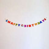 A Happy Birthday wool felt garland is displayed against a stark white backdrop, evoking a cheerful and celebratory atmosphere. This handcrafted felt garland spells "HAPPY BIRTHDAY" in vibrant rainbow letters: red, orange, yellow, green, blue, and purple. The string features whimsical party hats on each end and a delightful cake with a candle in the middle, enhancing its festive appeal.