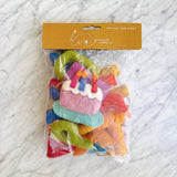 A packaged Happy Birthday felt garland rests on a marbled surface, ready for celebration. Inside its clear plastic bag, colorful felt figures are visible, including rainbow-toned alphabet letters, whimsical party hats, and a delightful cake with a candle. The kraft paper header on the packaging displays a brand logo and '100% wool / Made in Nepal'.