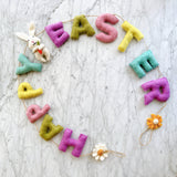 A festive Happy Easter felt garland is playfully arranged in a semi-circle on a white marble surface. This charming wool felt garland spells 'Happy Easter' in individual pastel pink, green, yellow, blue, and purple letters. It features a white felt bunny with long ears and black eyes holding a carrot on the left, a felt daisy at the bottom, and a felt sunflower on the right, all strung with jute thread.