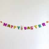 A vibrant wool felt Happy Easter garland hangs against a plain white wall, creating a cheerful and festive mood. It features individual pastel wool felt letters spelling 'Happy Easter' strung on a thin jute thread. Each end is adorned with a small wool felt daisy and sunflower. A white wool felt bunny clutching a tiny orange carrot is attached between the words, adding a playful touch.