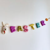 A pastel tone 'Happy Easter' wool felt garland is displayed against a clean white wall. The handmade garland spells out 'Happy Easter,' with each letter in a whimsical pastel color like purple, green, pink, yellow, and teal. It is strung on a jute string and adorned with a white felt bunny holding a felt carrot, a small orange felt sunflower, and felt daisies, adding a playful springtime charm.