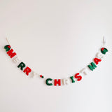 A felt Merry Christmas garland is hung against a plain white wall, suspended in a gentle arc. This hand-felted and stitched garland spells “Merry Christmas” with individual felt letters, alternating in classic red, white, and green colors. The letters are connected by a string, interspersed with small red, green, and white felt balls, evoking a festive and cheerful holiday charm.