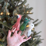 A handmade green wool felt gator finger puppet is playfully showcased by a female hand against a blurred, subtly lit Christmas tree backdrop. This unique felt toy for kids features a long body, a red smiling mouth, and big white eyes, highlighting its potential for creative interaction and festive fun.