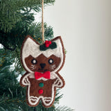 A needle felted gingerbread cat ornament hangs from a green Christmas tree, creating a joyful holiday atmosphere. The light brown cat-shaped ornament features white felt outlining, mimicking gingerbread cookie icing, with a white 'frosting' on its head. It has a red and green holly accent, stitched black eyes, mouth, white whiskers, a red bow tie, and small red and green felt dots on its body.
