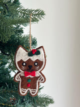 A charming wool felt gingerbread cat ornament hangs from a lush green Christmas tree. This handmade felt cat ornament features white 'frosting' along its head and sides, a red bow tie, small round eyes, and stitched whiskers. Its body is adorned with small red and green wool felt circles, suspended by a jute string.