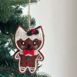 A charming wool felt gingerbread cat ornament hangs from a lush green Christmas tree. This handmade felt cat ornament features white 'frosting' along its head and sides, a red bow tie, small round eyes, and stitched whiskers. Its body is adorned with small red and green wool felt circles, suspended by a jute string.