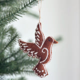 A handcrafted felt gingerbread dove ornament hangs gracefully from a green Christmas tree branch, evoking a cozy holiday atmosphere. The charming brown felt dove features white felt accents that resemble frosting on a gingerbread cookie, highlighting its whimsical design.