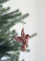 A felt gingerbread dove ornament, crafted from textured brown felt with needle felting details, hangs festively on a Christmas tree. This handmade Christmas ornament features sweet white accents mimicking icing and is suspended by a jute string, adding a joyful charm to the greenery.