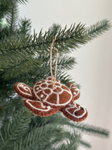 A warm brown wool felt turtle ornament, part of the Felt Gingerbread Collection, hangs on a festive green Christmas tree. This unique Christmas ornament features white decorative stitching in geometric patterns on its shell, head, and flippers, resembling a gingerbread cookie. Small black bead eyes and a jute string for hanging complete the design.