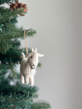 A charming needle felted goat Christmas ornament, crafted from light-colored wool felt, hangs from a green Christmas tree branch. This handmade felt goat ornament features a textured, hand-stitched surface with small ears, a short tail, and a gentle stitched smile. A jute string is attached at the top for easy display.
