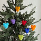 A set of 7 rainbow felt heart ornaments adorns a vibrant Christmas tree, presented against a plain white background. These handmade felt ornaments feature beautifully shaped hearts in yellow, purple, red, orange, blue, and green. Each colorful felt ornament includes twine for easy hanging, bringing a joyful and festive atmosphere to the tree.