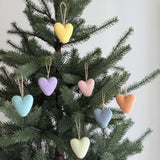 Seven wool felt heart ornaments from a rainbow set are closely displayed on a lush Christmas tree. Each unique handmade felt ornament showcases a soft pastel hue, including yellow, lavender, light pink, light blue, light gray blue, peach, and light green, suspended by rustic jute string. The handcrafted felt ornaments create a festive yet minimalist display, their soft texture contrasting with the tree's smooth needles.