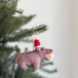 A light purple wool felt hippo ornament, featuring a chunky, needle-felted design, hangs from a Christmas tree branch. This cheerful hippo ornament wears a small red Santa hat with a white pompom and has visible white teeth. A jute string attaches the unique Christmas ornament to the festive branch.