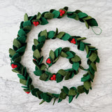 A wool felt holly garland is arranged in a spiral on a white marble surface, creating a festive display. The handcrafted garland features alternating pairs of dark green and light green holly leaves. Each leaf pair is adorned with clusters of three small red berries, all strung on a green jute thread.