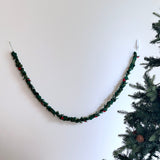 A wool felt holly garland hangs on a white wall next to a Christmas tree, creating a festive atmosphere. The garland features alternating dark green whimsical holly leaves and small red berries strung along a jute thread, forming a cheerful pattern.