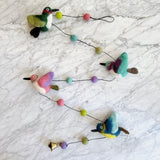 A vibrant felt hummingbird garland is whimsically arranged on a marbled surface. This cheerful felt garland features four colorful felt hummingbirds in green, pink, blue, and teal hues, strung along a thin black cord. Colorful felt balls are interspersed between the birds, and a small copper bell hangs at one end.