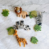 A handcrafted felted wool jungle animal garland is displayed on a white marble surface, presenting a playful and natural aesthetic. It features a tan lion with a dark brown mane, a gray elephant, a black and white striped zebra, and an orange giraffe with brown spots. Green monstera leaves are interspersed among the animals, all connected by a rustic jute thread.