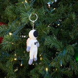 A playful wool felt astronaut keychain hangs from a branch of a Christmas tree adorned with tiny lights. The handmade felt keychain features a white spacesuit, a black visor, and gray accents on its hands and feet, adding a whimsical touch to the festive holiday decor. It is attached to a metal keychain.