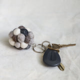 A handmade ball pompom felt keychain is displayed on a softly textured white surface, alongside a modern car key and a traditional brass key. This unique wool felt keychain features an array of small, tactile felt balls in neutral shades of white, grays, and browns. Its handcrafted appearance highlights a simple, functional aesthetic.