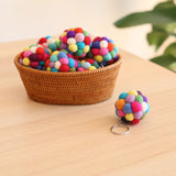 Ball pompom felt keychains are displayed on a light wooden table with a subtle green backdrop. One vibrant, spherical wool felt ball pompom keychain, featuring a metal key ring, sits beside a woven basket filled with more colorful felt balls in shades like red, blue, and green.