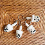 Four handmade wool felt Boba Tea keychains are displayed on a light brown woven surface, showcasing their unique charm. Each felted keychain features a rounded beige and black boba tea cup, complete with hand-stitched details, a fluffy white straw, and a silver metal key ring.