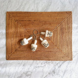 Wool felt boba tea keychains are arranged on a brown woven mat against a white marble background. Each cylindrical keychain features a white foam-like top, a tan body, and small black hand-stitched tapioca pearls with a tiny straw. A metal clasp keychain is attached to each charming felt boba tea design.